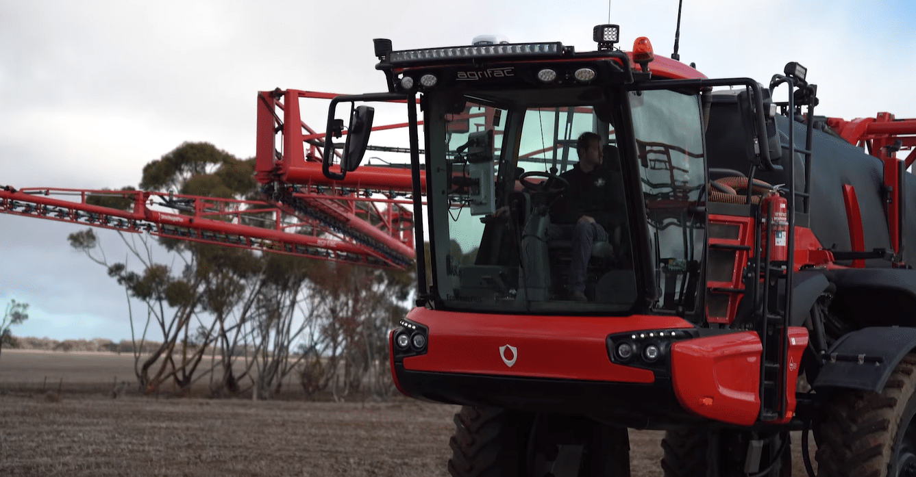 Red self-propelled sprayer tractor with extended boom in a field, cab facing forward and driver inside.