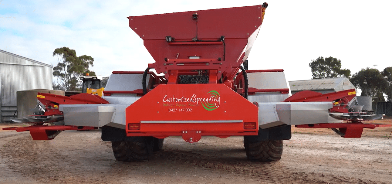 Rear view of a large red farm spreader with a raised hopper and extended side arms; 'Customized Spreading' logo on the back.