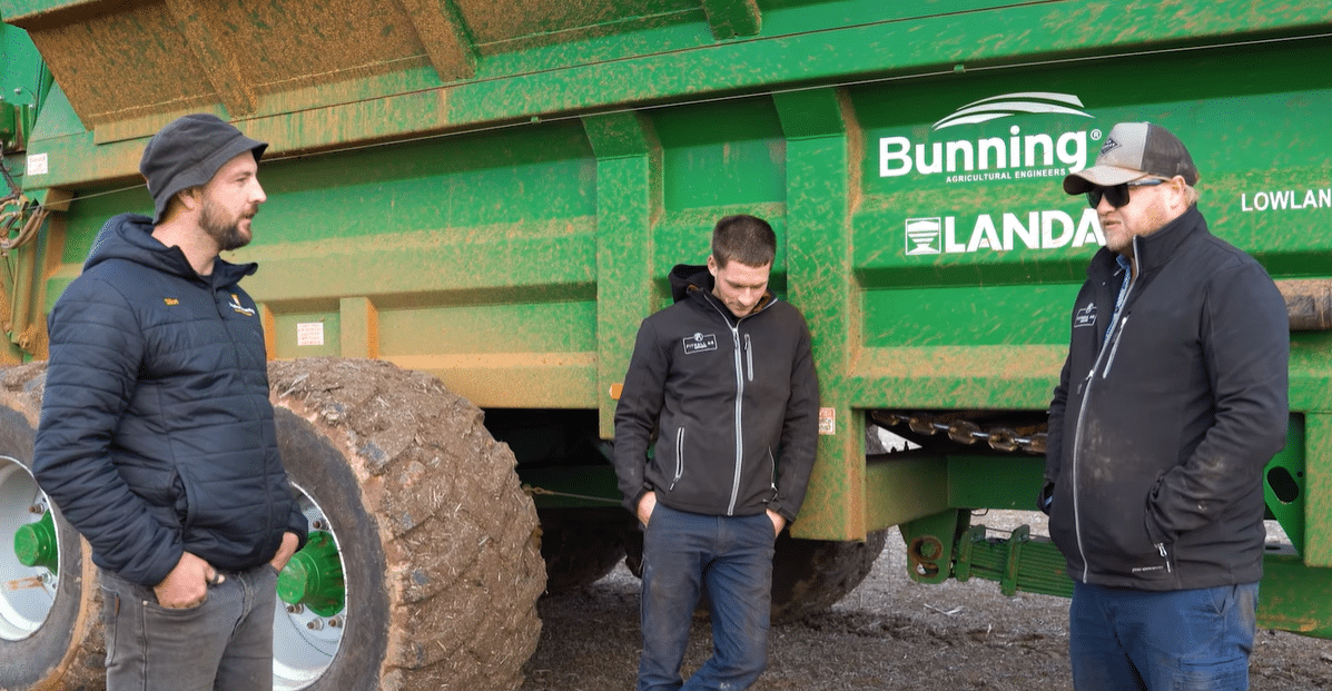 Three men in jackets stand beside a large green agricultural trailer with muddy tires, talking outdoors.