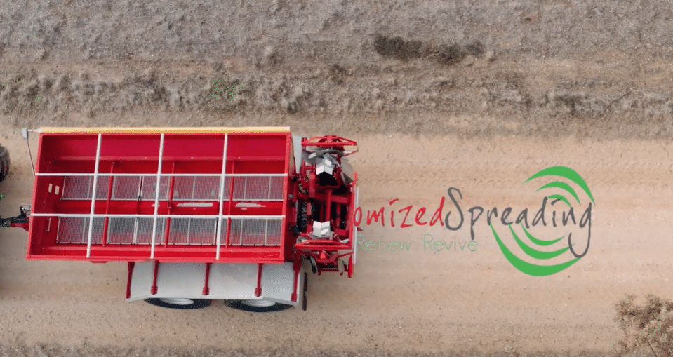 Top-down aerial view of a red agricultural spreader attached to a truck driving along a dirt road, with a green 'Customized Spreading' logo nearby.