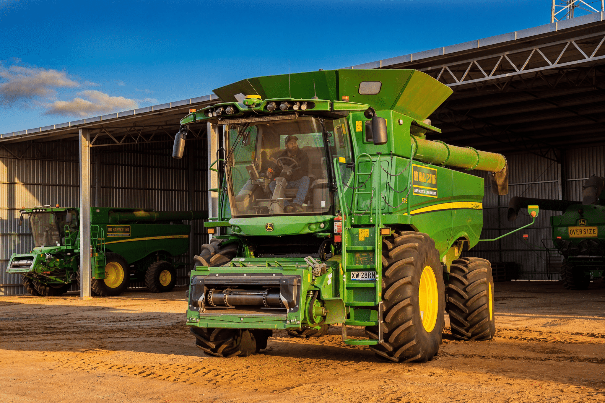 Green John Deere combine harvester parked under a metal shed with a blue sky above; another machine visible in the background.