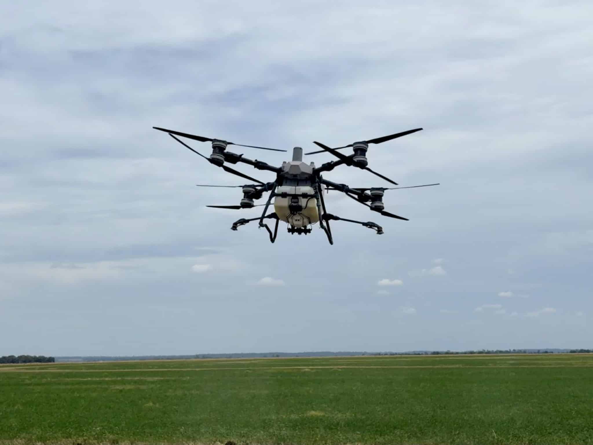 Large multi-rotor drone in flight over a grassy field with a cloudy sky, view from below of its rotors and landing gear