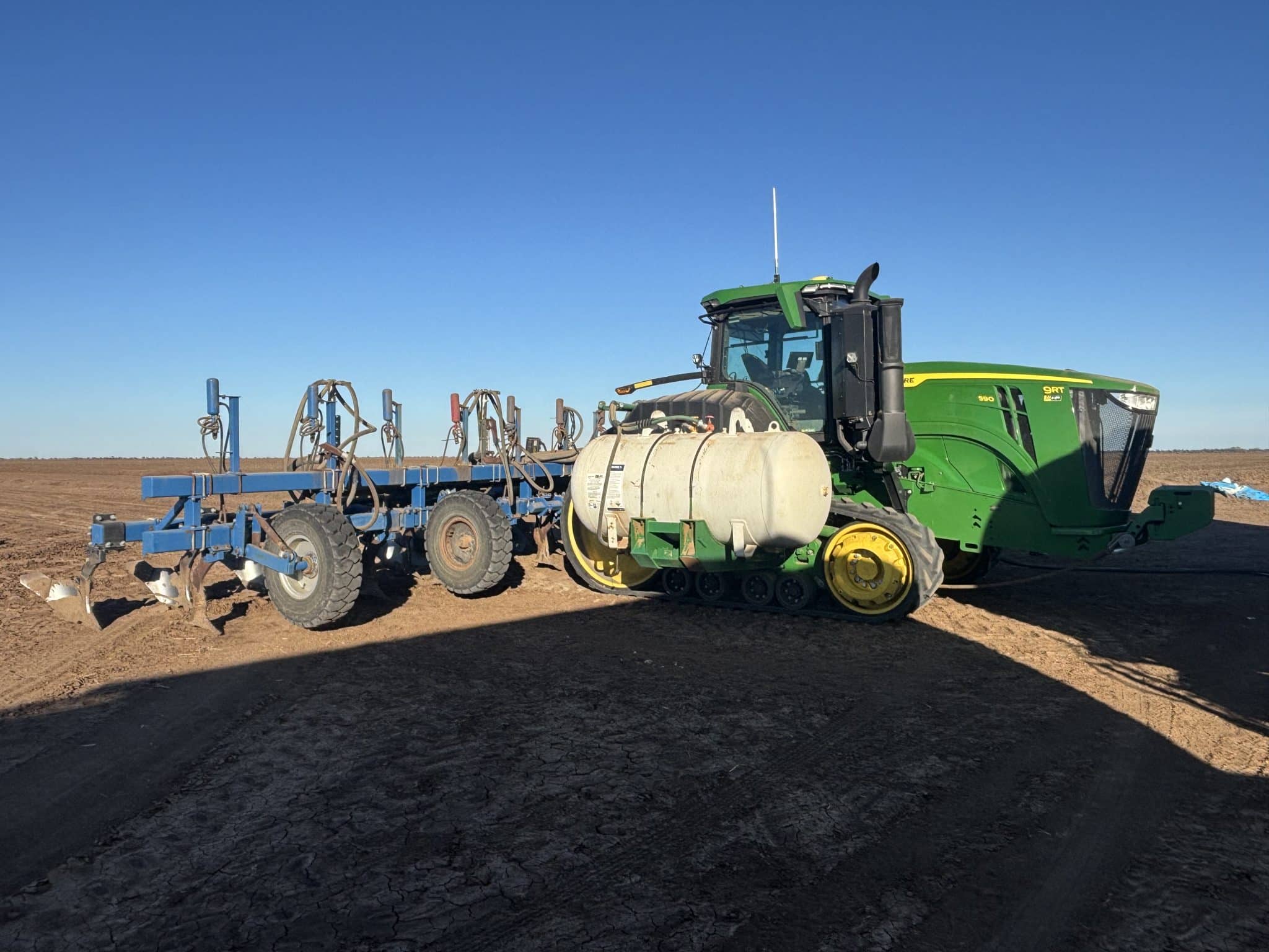 Green tractor attached to a blue farming implement on a dry, cracked field beneath a clear blue sky.