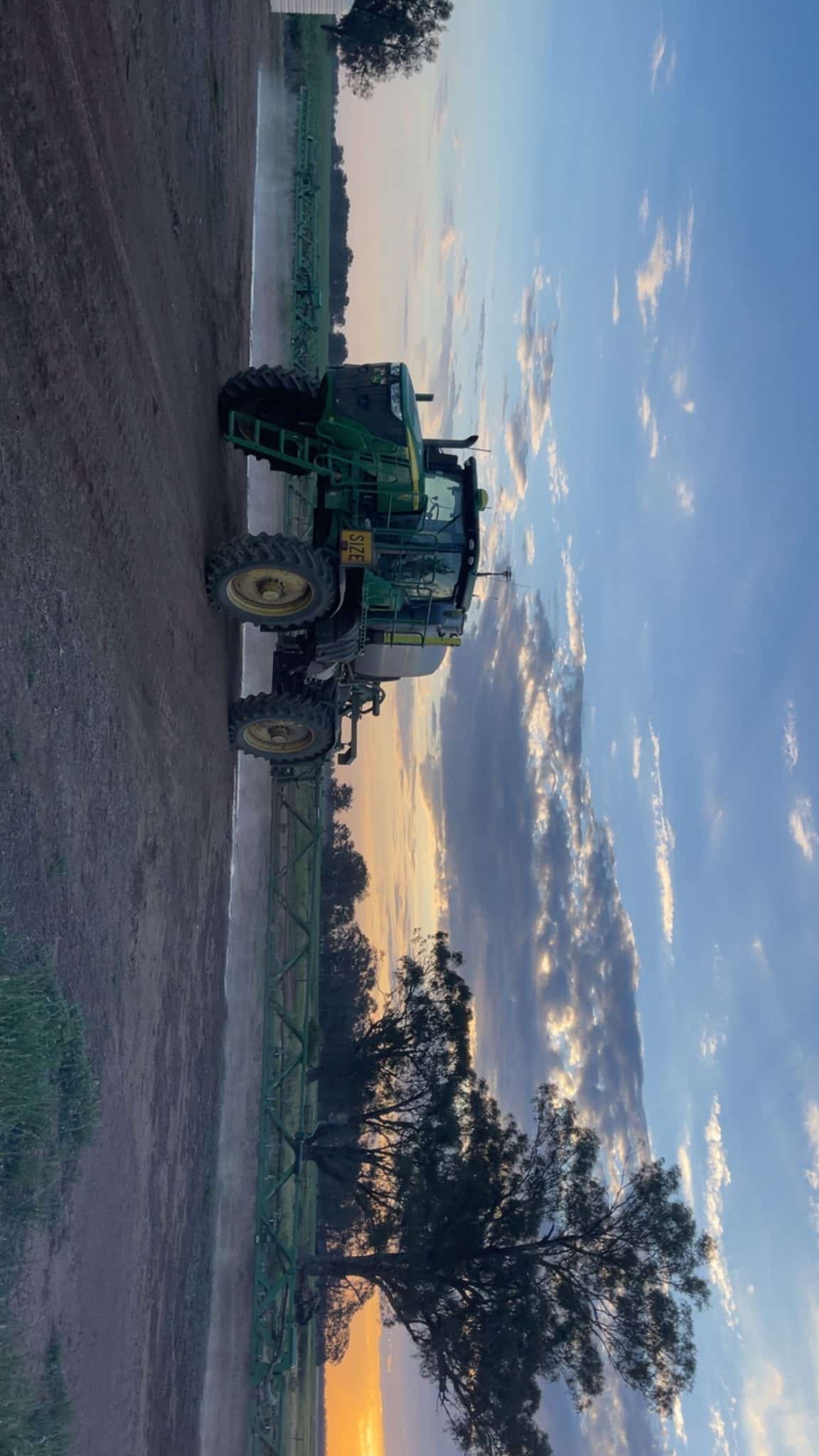 Green farming tractor with header attached, parked in a plowed field at sunset.