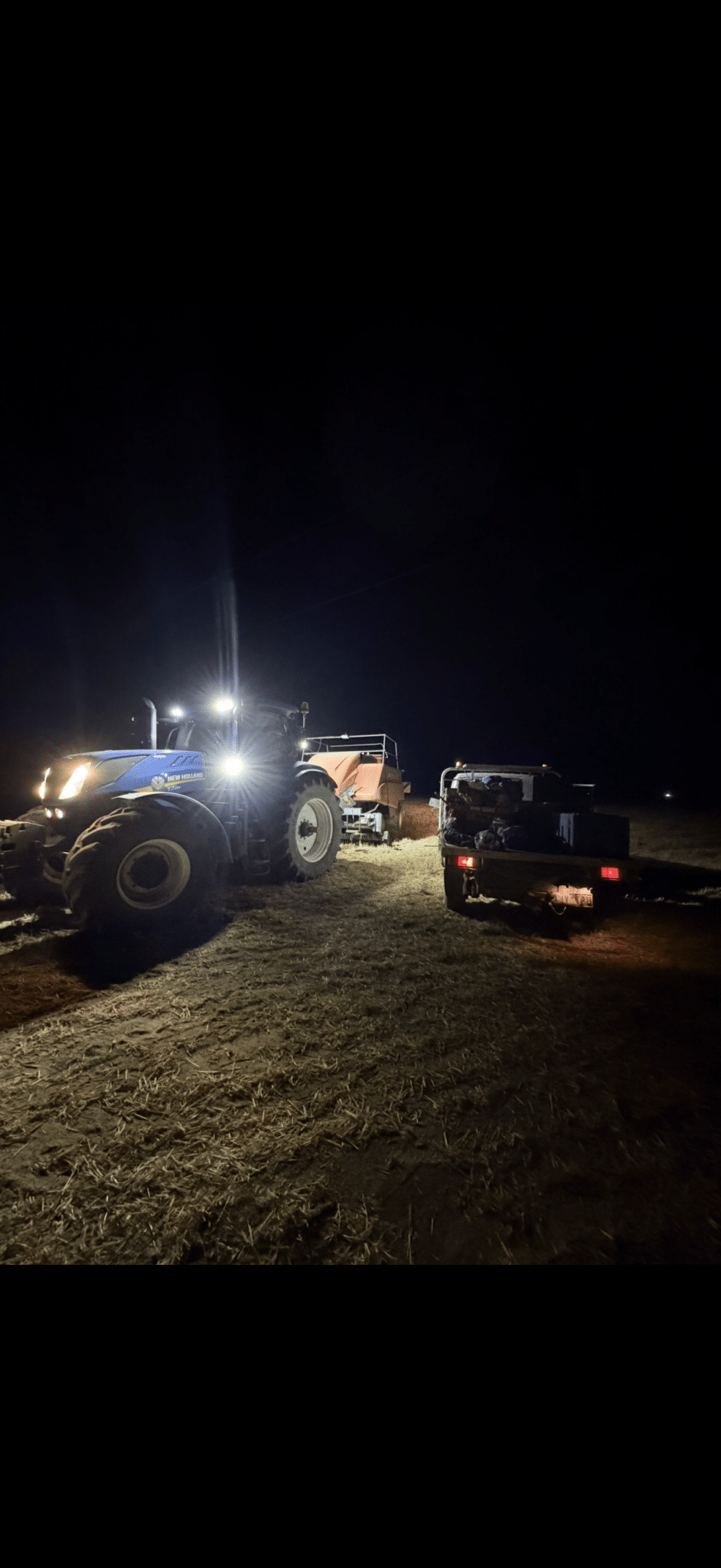 Blue New Holland tractor with bright headlights at night, connected to a baler and trailer in a field shiftlighted by work lamps in the foreground.