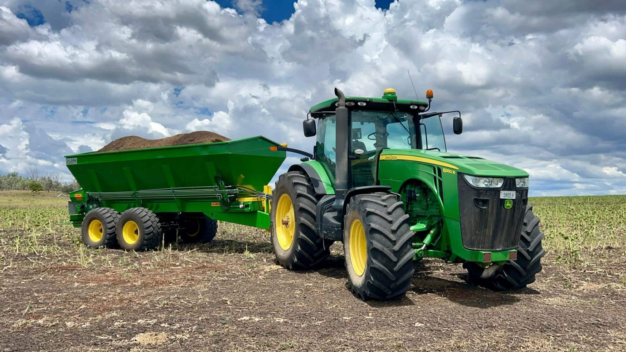 Green John Deere tractor with a matching green grain cart attached, parked in a harvested field under a cloudy sky.