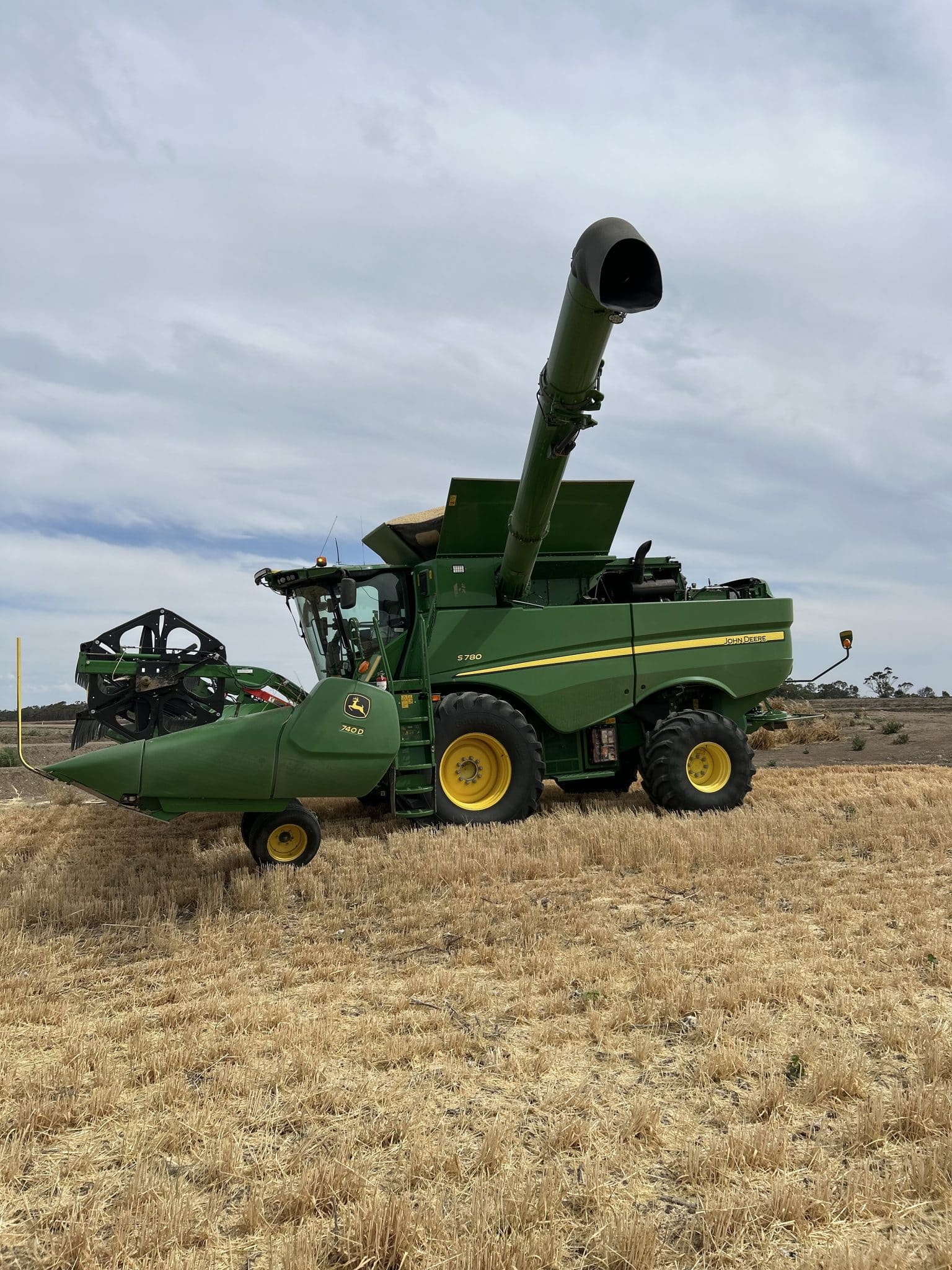 John Deere combine harvester with a raised unloading auger in a harvested field under a cloudy sky