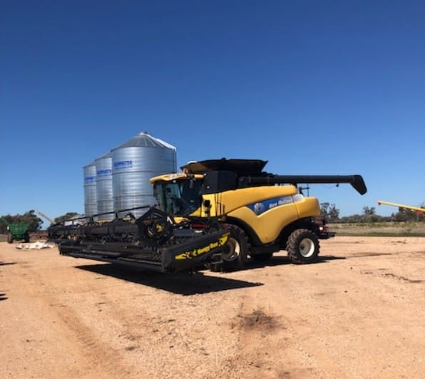 Yellow combine harvester with header attachment on a dirt lot, blue sky and grain silos in the background.
