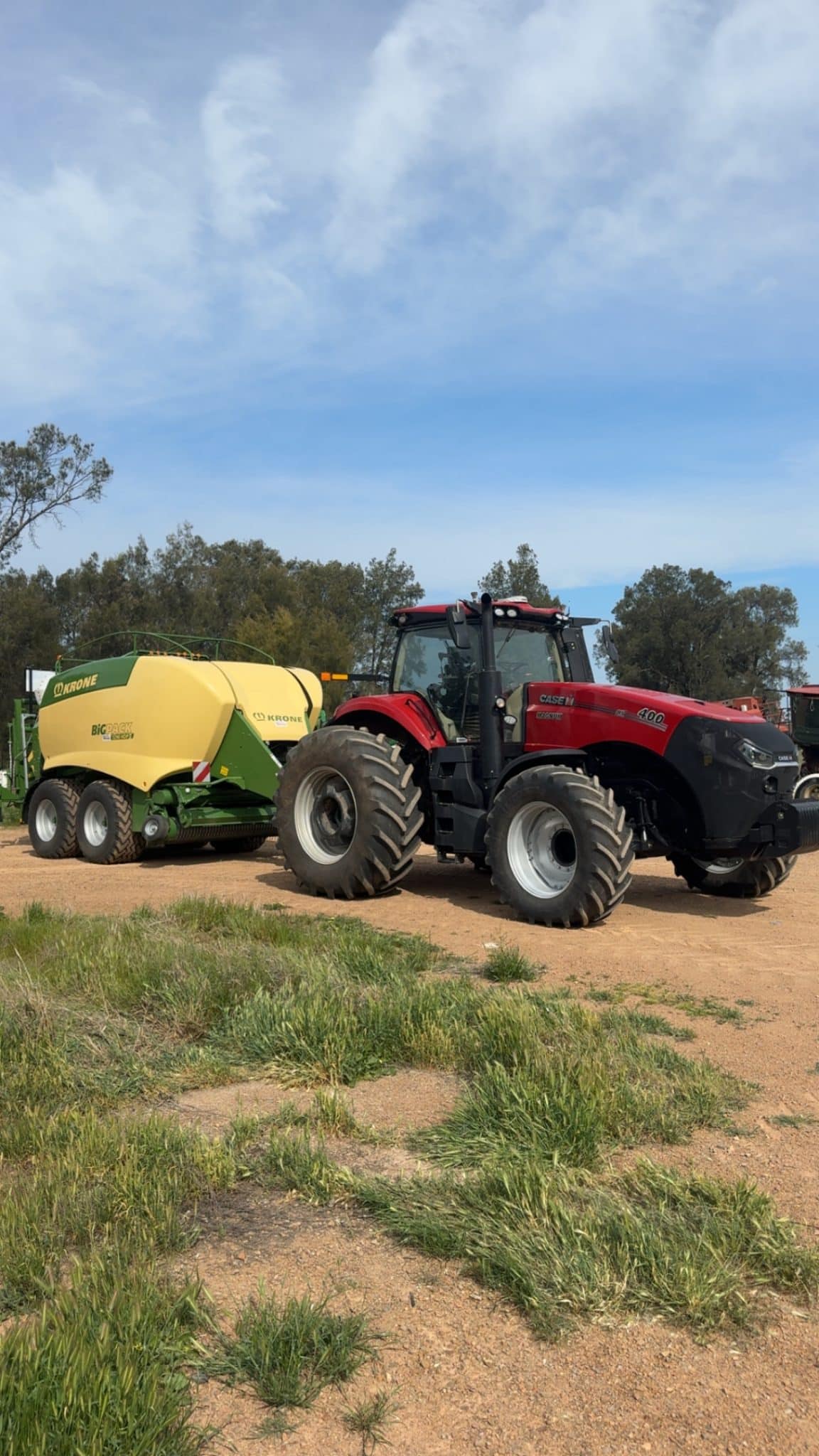 Red Case IH tractor with large off-road tires connected to a yellow Krone round baler on a dirt field. Trees in the background under a partly cloudy sky.