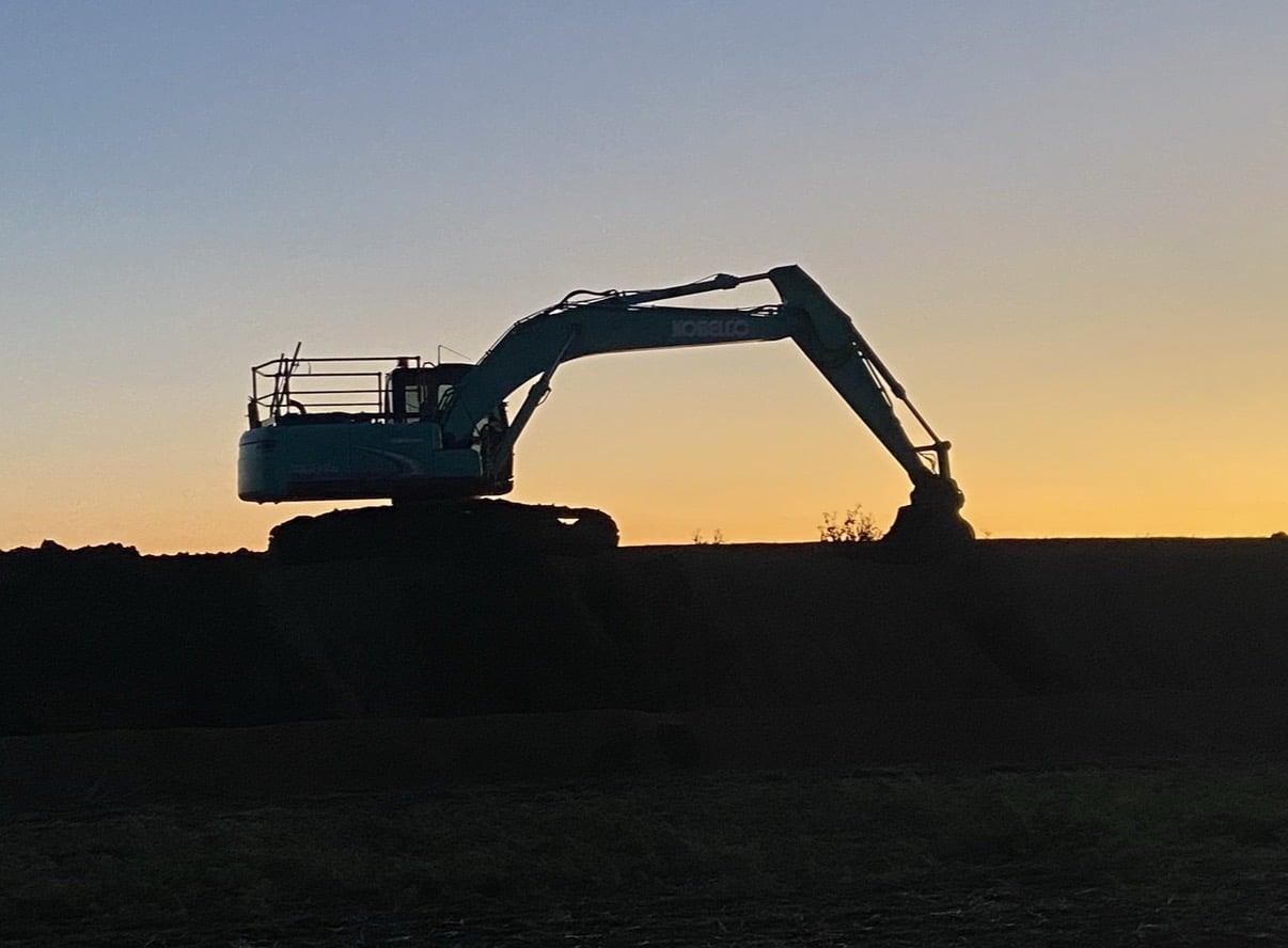 Silhouette of a large excavator on a dirt mound at sunset with an orange sky behind it.",