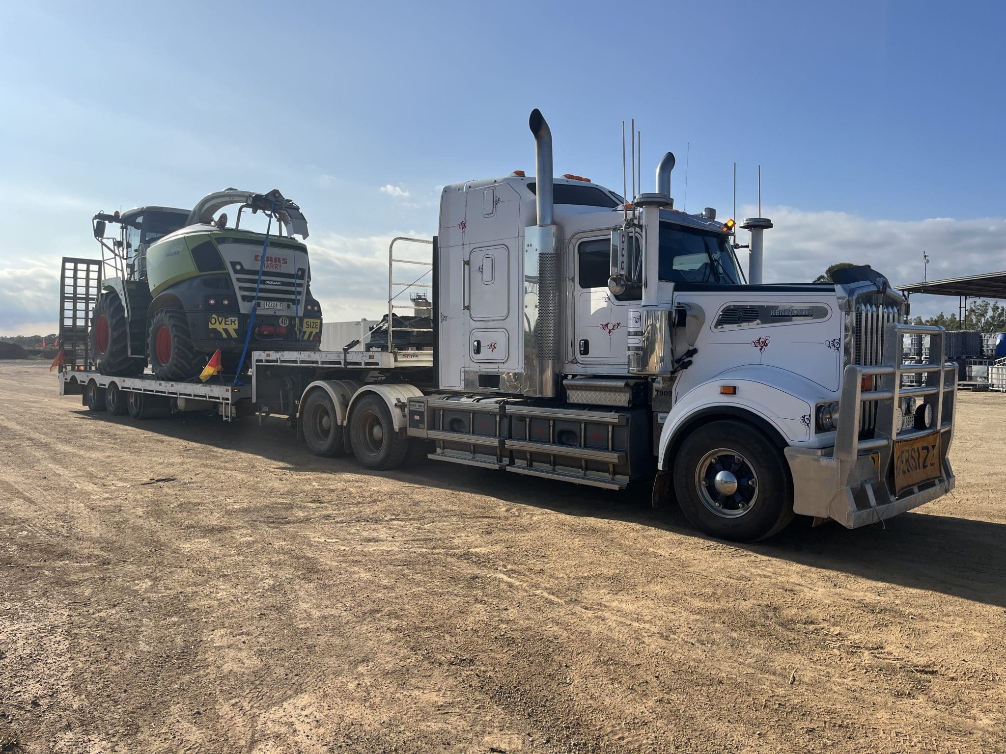 White semi-truck hauling a flatbed trailer with a green CLAAS Jaguar forage harvester in a dusty yard under a blue sky.