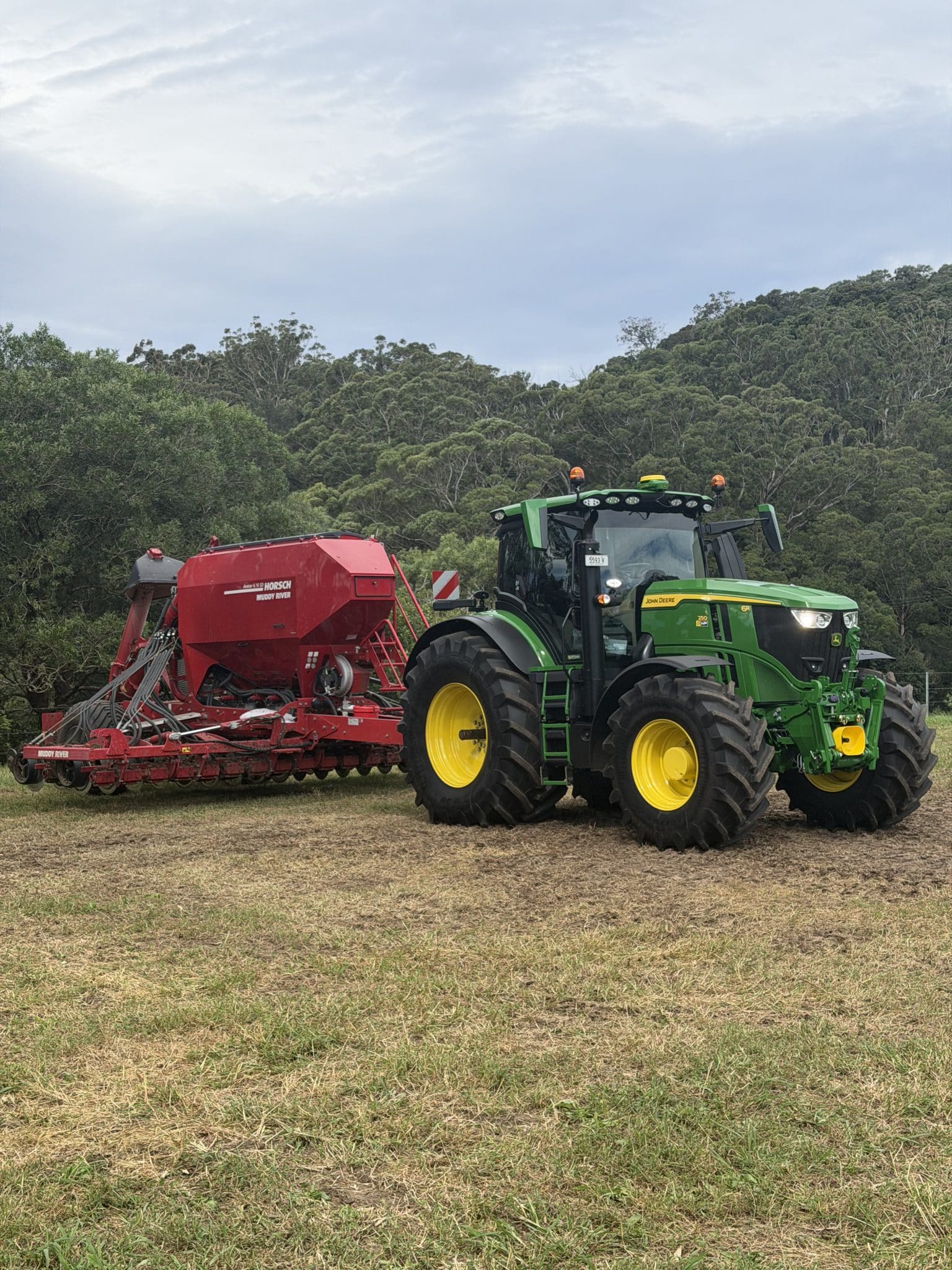 Green John Deere tractor with yellow wheels paired with a red farm implement in a dry field, with a tree line in the background.