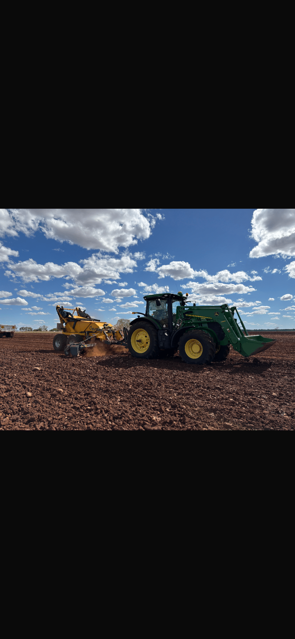 Green tractor with a yellow tillage implement working a brown, freshly plowed field under a bright blue sky with scattered clouds.