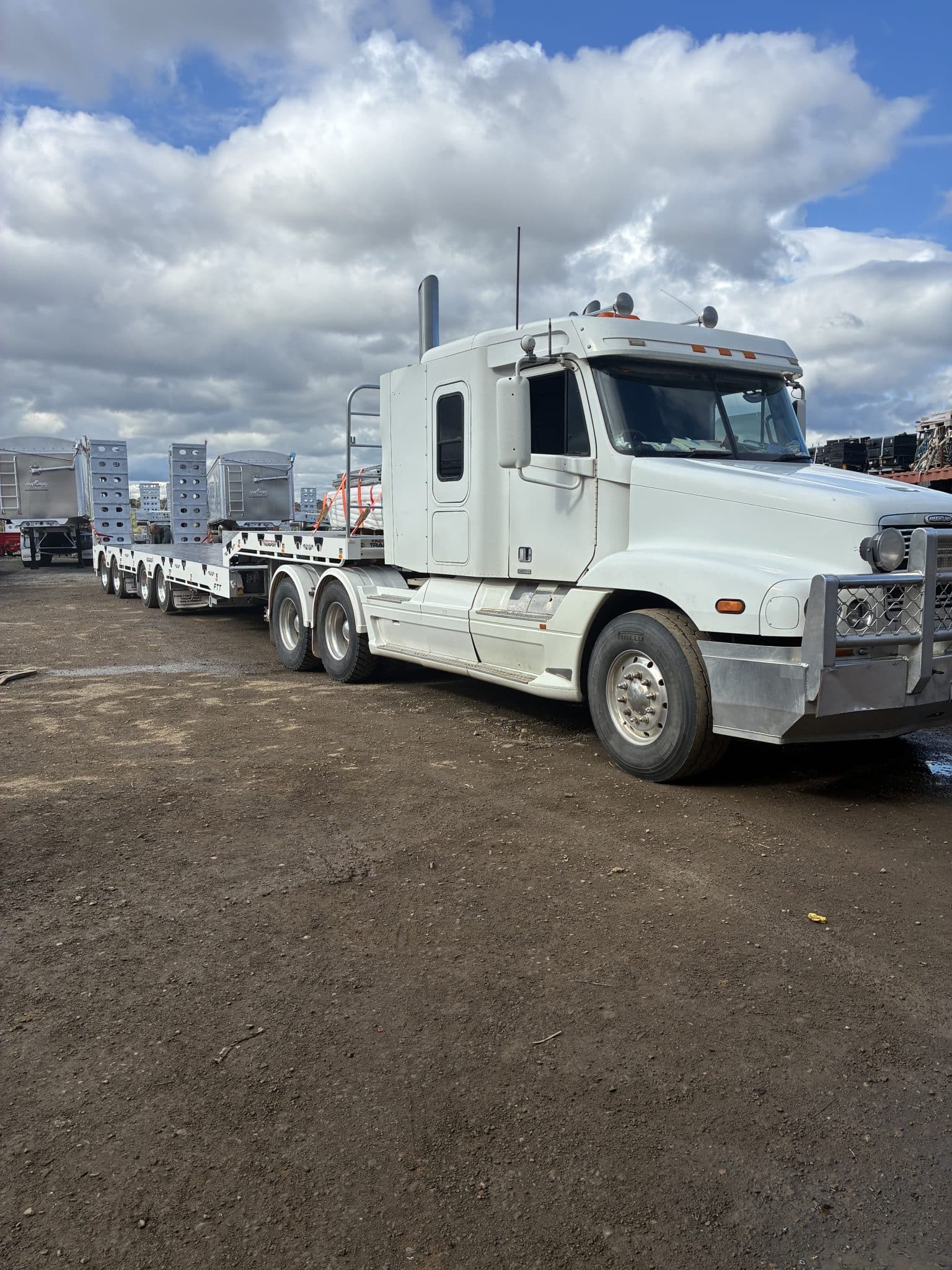 White semi-truck tractor with a long flatbed trailer carrying metal components in an industrial yard under a cloudy sky.