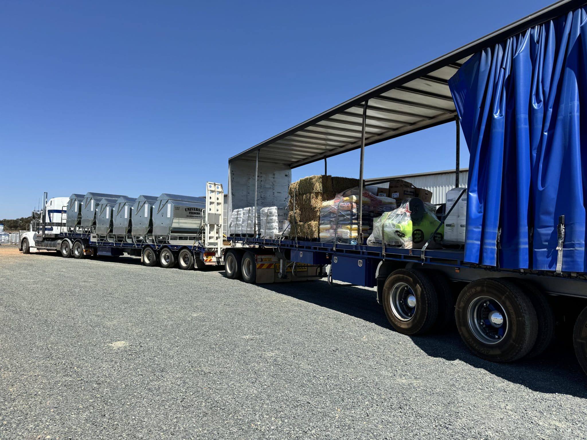 Long flatbed truck trailer with a blue curtain side open, carrying hay bales and pallets under a metal shelter against a clear blue sky.