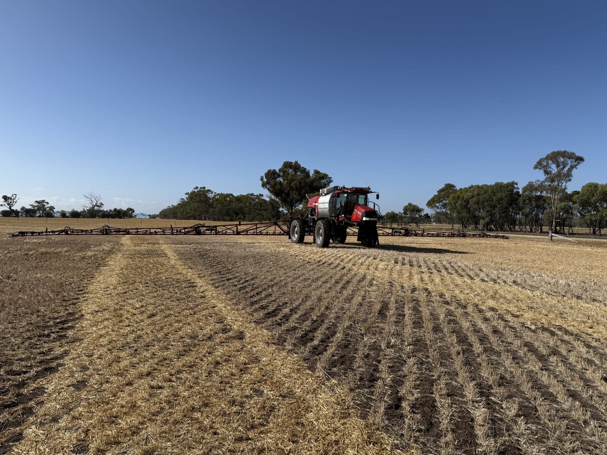 Red farming tractor beside a long irrigation boom in a dry, harvested field under a clear blue sky.