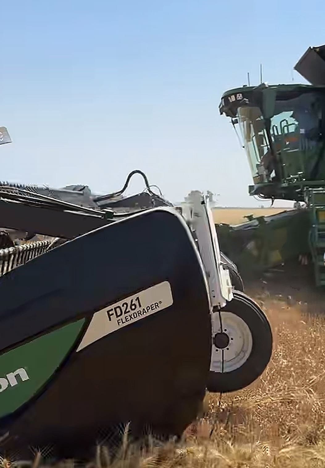 Harvester mower in a field under a clear blue sky, showing a labeled FD261 FLEXDRAPER component with a green and black body and a large wheel in the foreground.