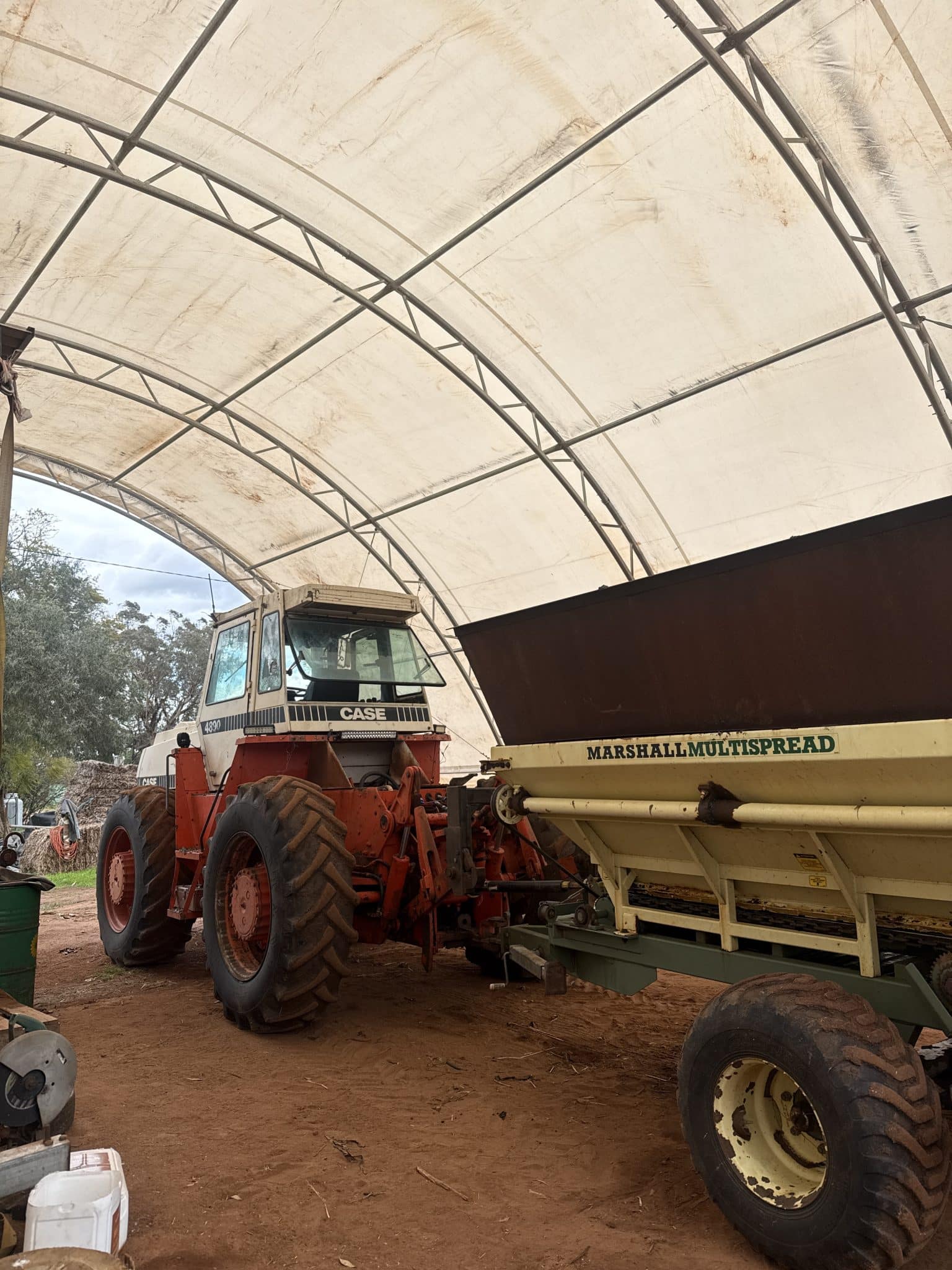 Open-air workshop with a large beige fabric-covered arch structure and orange CASE tractor parked beside a Marshall Multispread fertilizer spreader under the canopy, on a dirt ground.