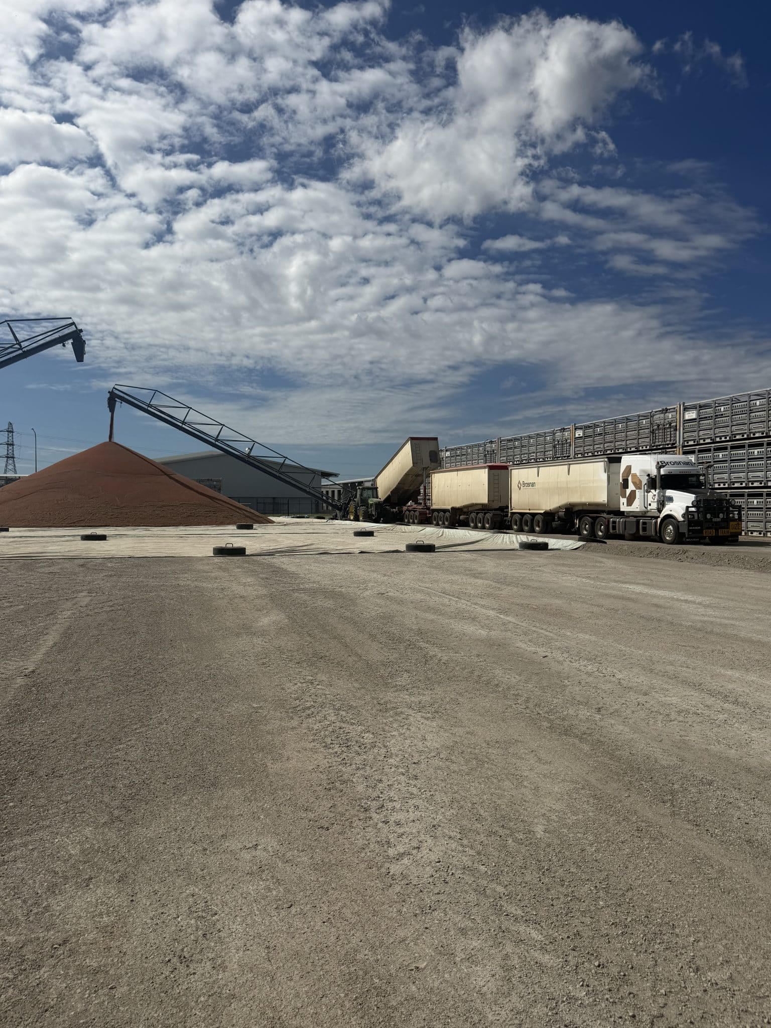 Industrial yard with a conveyor system and a large pile of material (sand/aggregate) beside trucks and trailers under a cloudy sky.