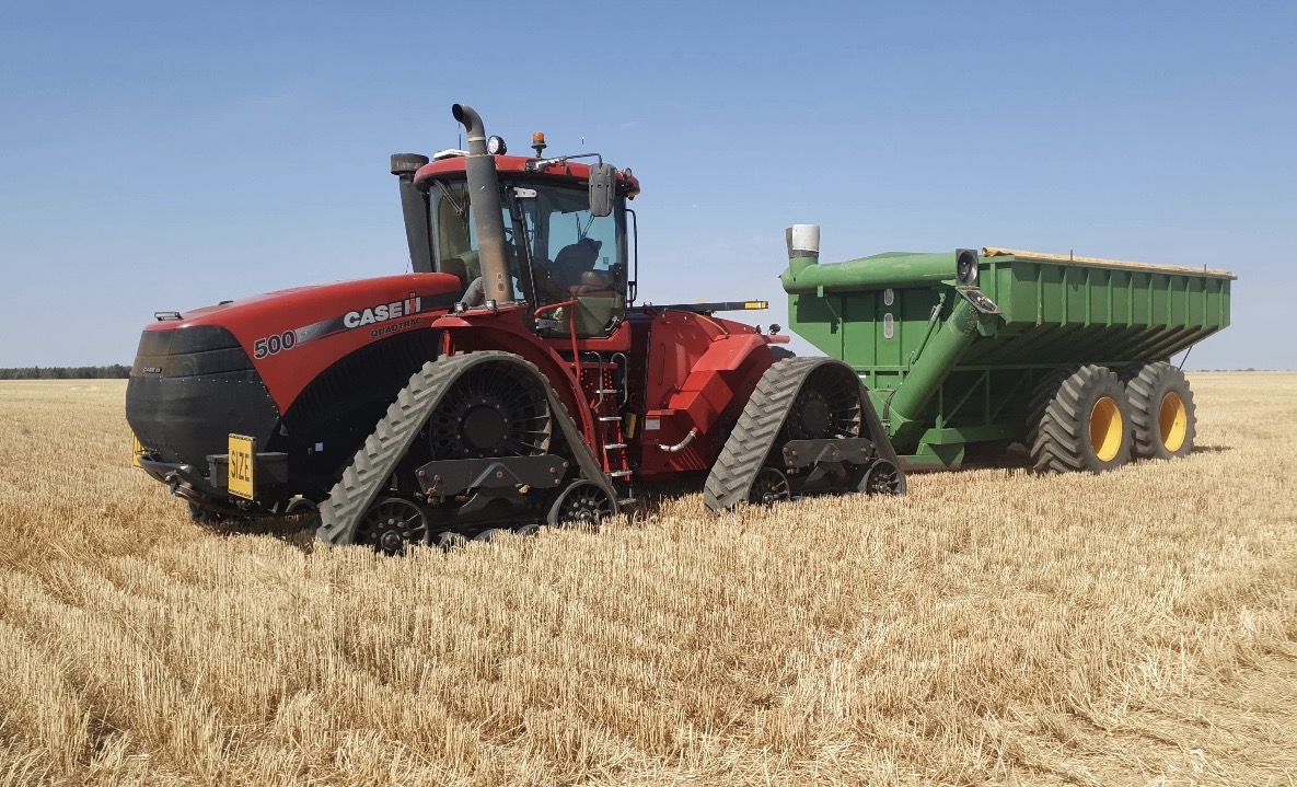 Red Case IH quad-track tractor pulling a green grain cart across a harvested wheat field under a blue sky
