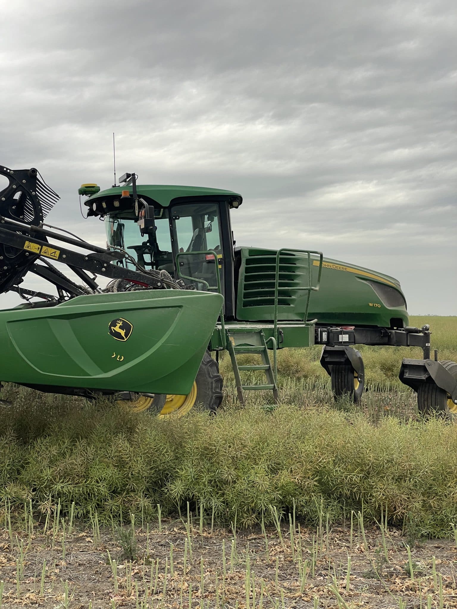 Green John Deere combine harvester with header attached, standing in a field under a cloudy sky; crops in foreground and stubble beneath.