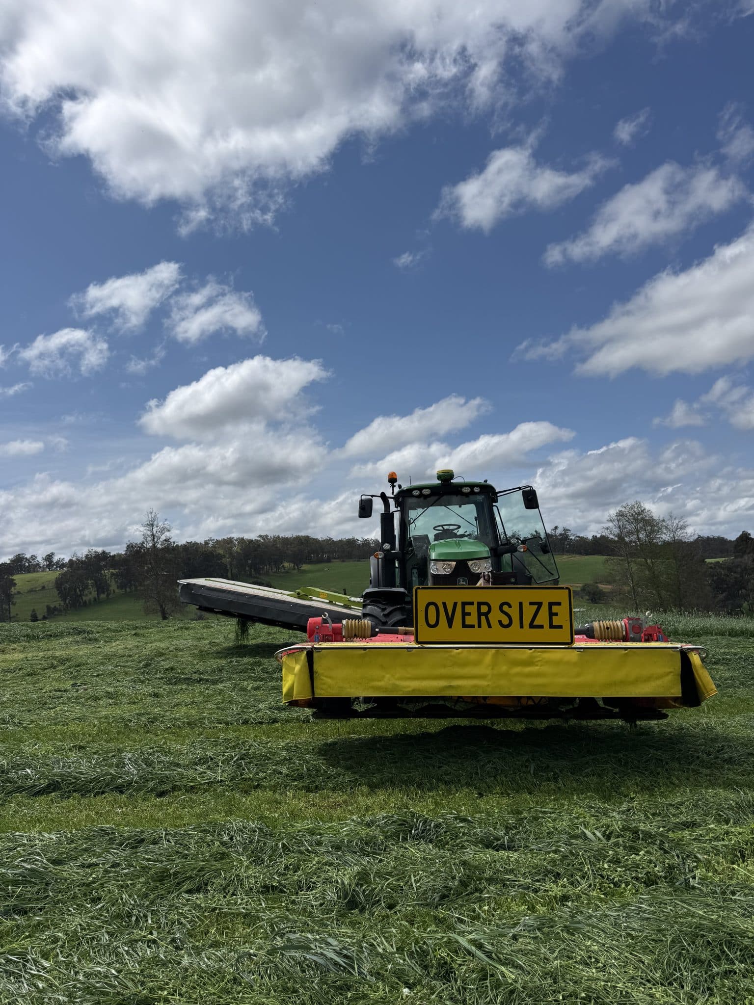 Green tractor with a yellow 'OVERSIZE' sign on the front, in a grassy field under a blue sky with scattered clouds.