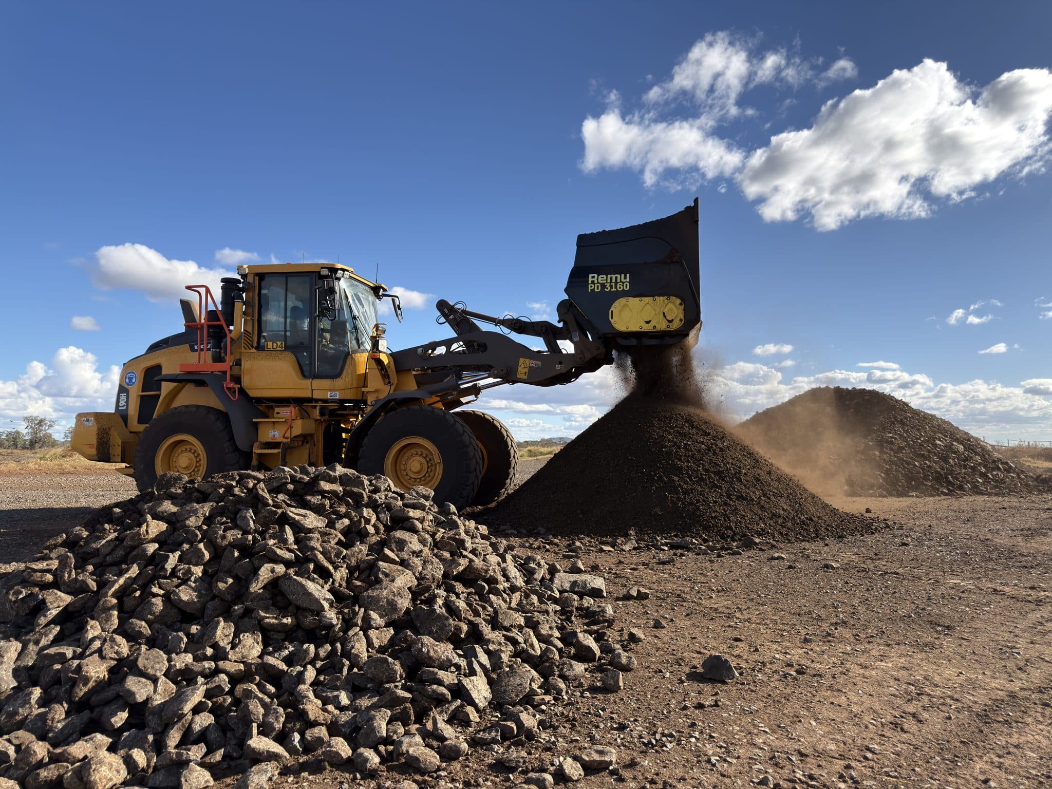 Large yellow construction loader unloading a pile of rocks against a bright blue sky.