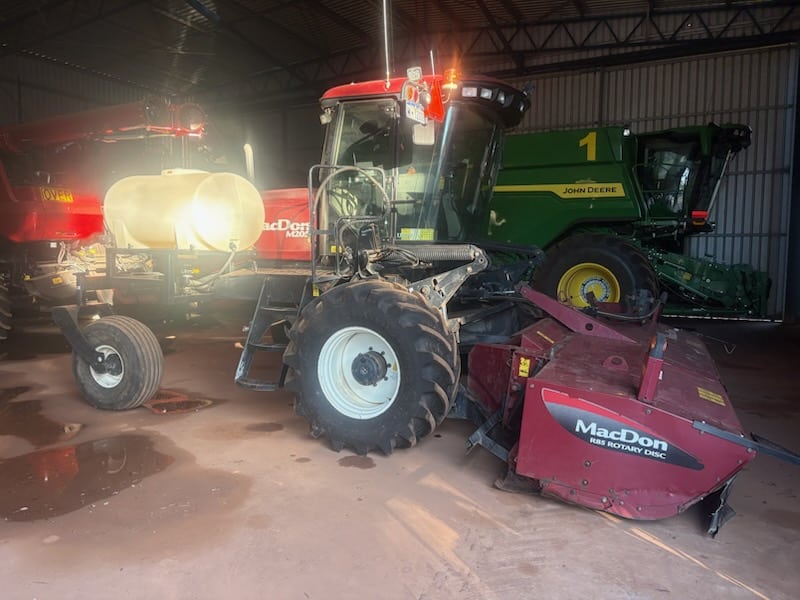 Inside a shed with farm equipment: a red MacDon rotary disc header hitched to a tractor, next to a green John Deere combine harvester in the background.