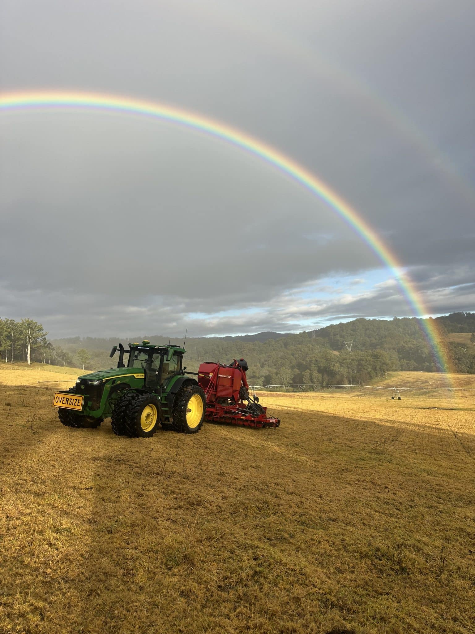 Green tractor with a red attachment sits in a dry field under a large double rainbow across a cloudy sky in a rural landscape
