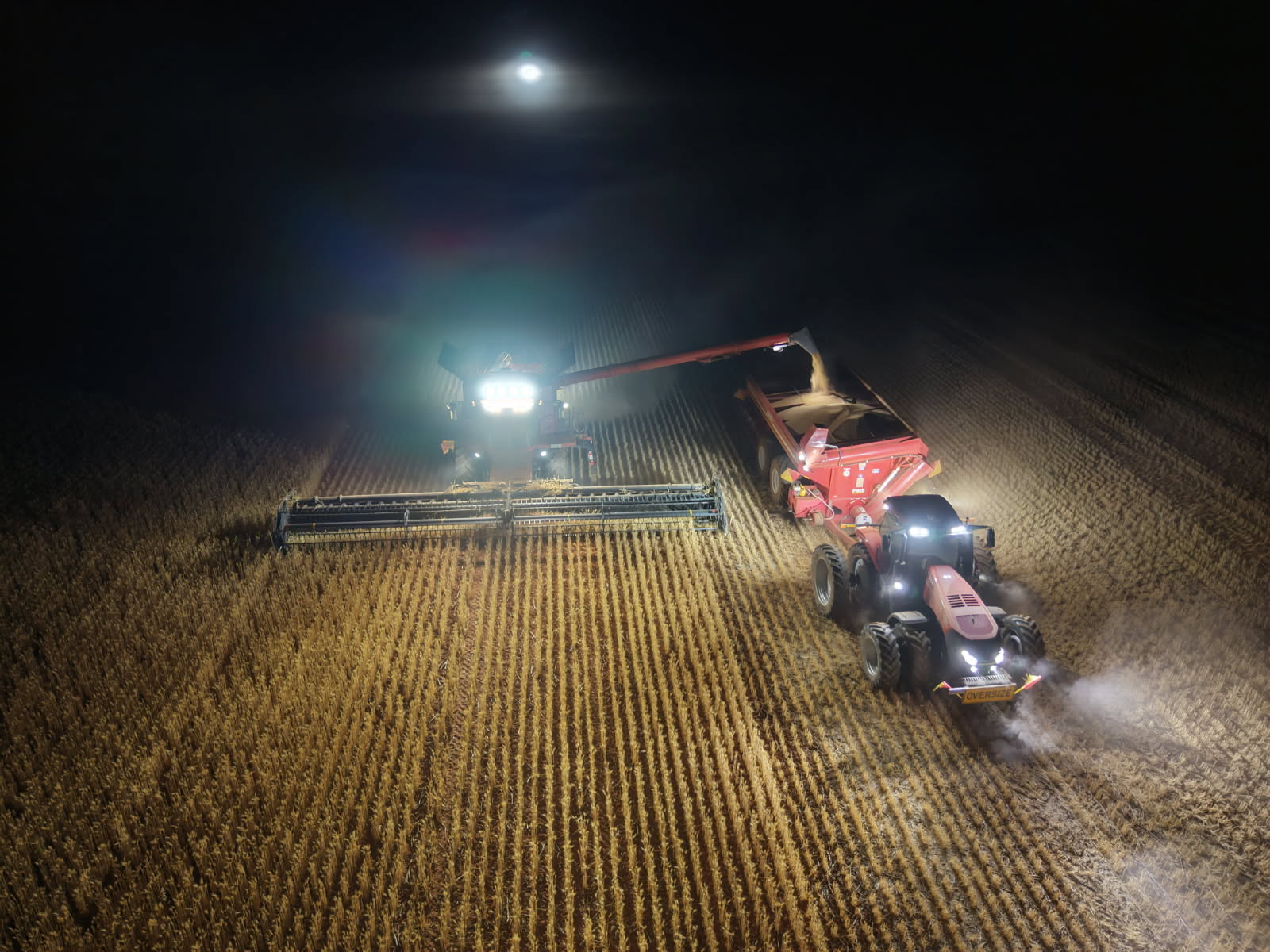 Nighttime farming scene with a combine harvester unloading grain into a red grain trailer under bright tractor headlights